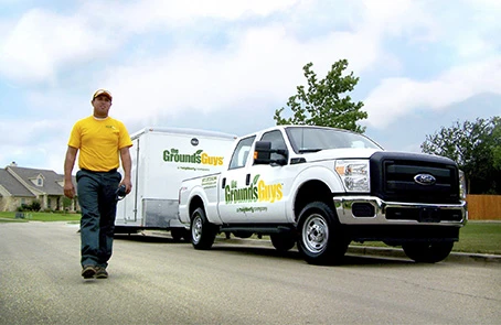 Grounds Guys worker in yellow branded T-shirt walking next to parked company truck and trailer.
