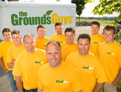 The Grounds Guys employees wearing branded company T-shirts smiling in front of company trailer.