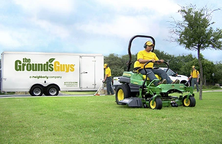Employee in Grounds Guys shirt using riding mower to cut grass, with company trailer in background.