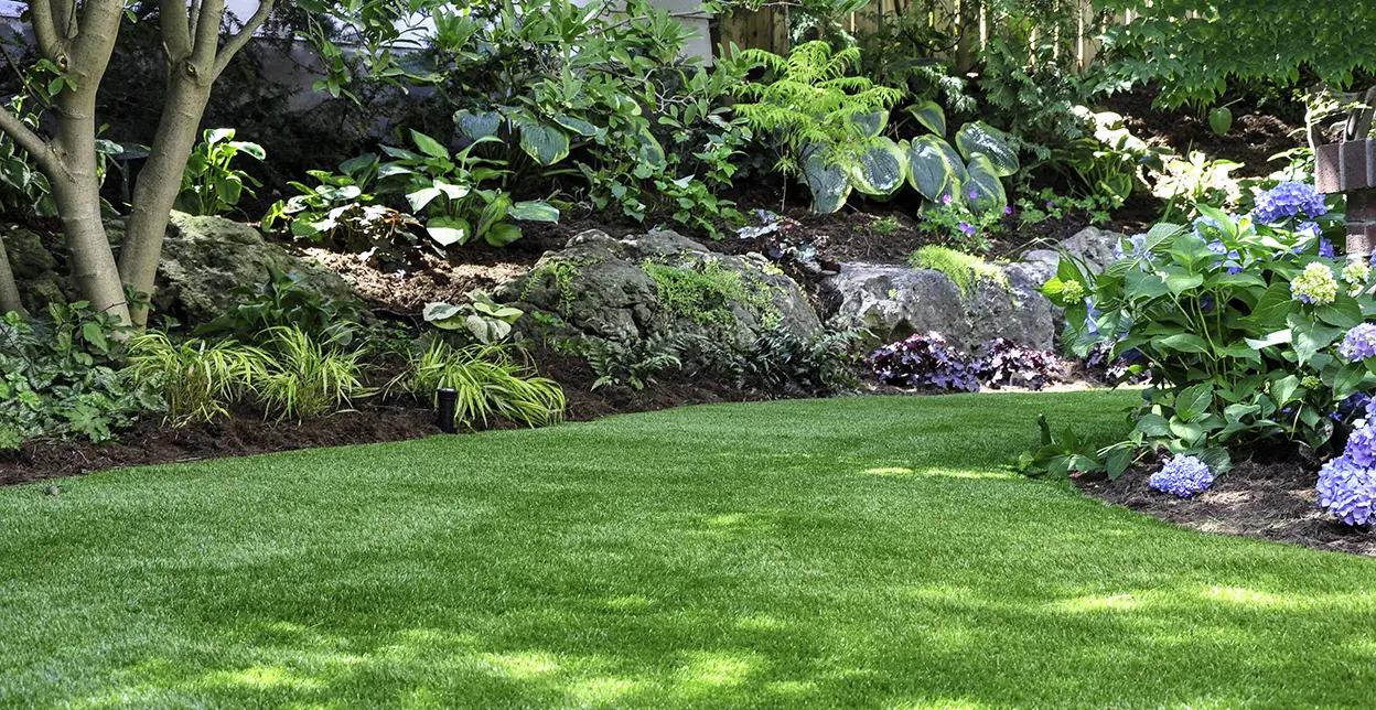 Artificial grass installed on a property's lawn, with lush flower beds along its edges.