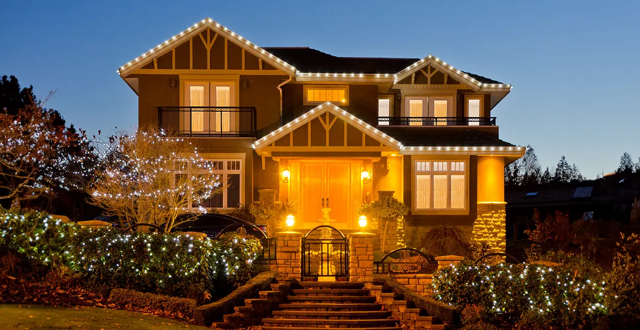 The front of a large home, with holiday lighting along its eaves and in its trees and bushes.