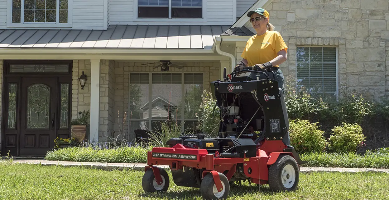 The Grounds Guys service professional aerating a home's front lawn on a stand-on aerator.