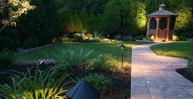 A well-landscaped backyard at night, with brick path and gazebo, illuminated by lighting.