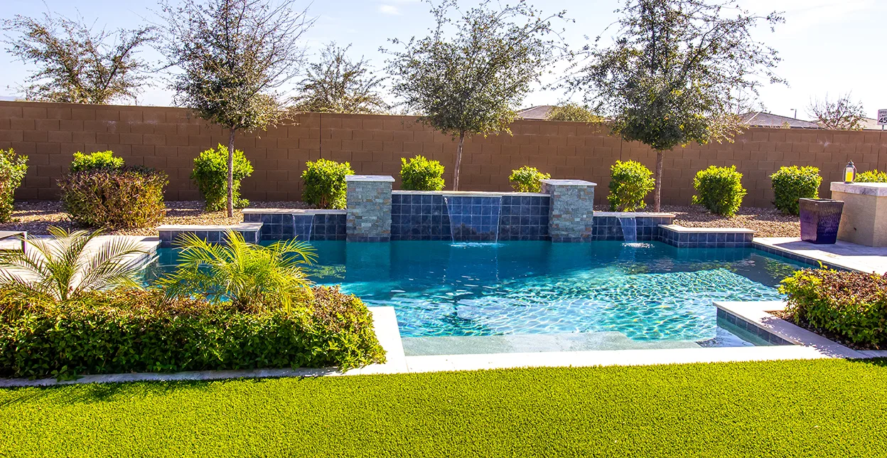 A backyard swimming pool with built-in waterfalls, bordered by shrubs and trees.