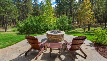 A stone firepit with Adirondack chairs facing a wooded backyard.