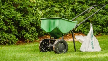 A fertilizer spreader on a lawn with bag of fertilizer next to it.