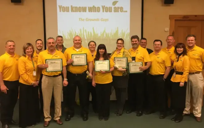 The Grounds Guys of Avon team holding certificates in front of a projection screen displaying The Grounds Guys logo.