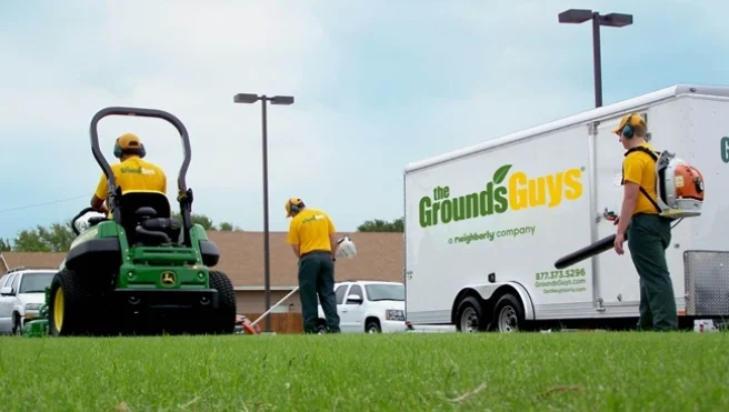 Three Grounds Guys service professionals performing lawn cleanup next to a branded company trailer.