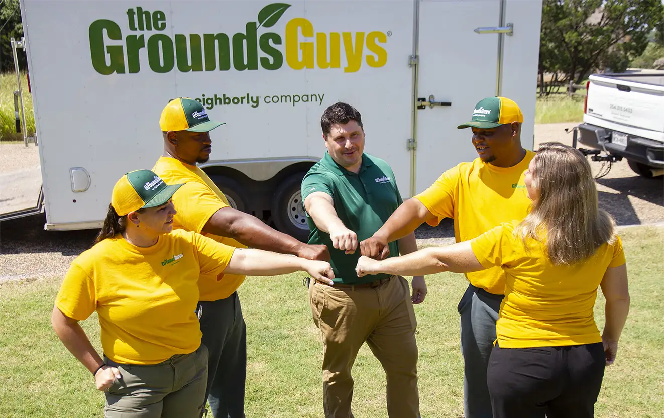 The Grounds Guys crew standing in a circle with their hands in the center, in front of a branded company trailer.