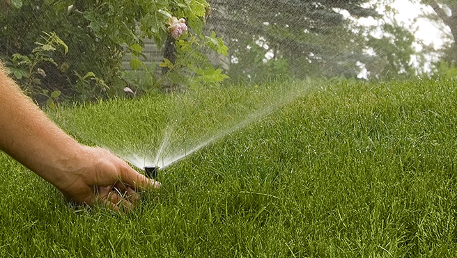 A service professional's hand adjusting a sprinkler head in grass as it sprays water.