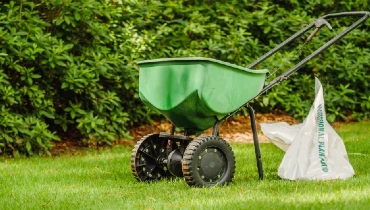 Manual fertilizer spreader and bag of fertilizer sitting on the green lawn.